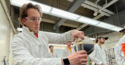 Student in a lab coat pouring a solution into a vessel.