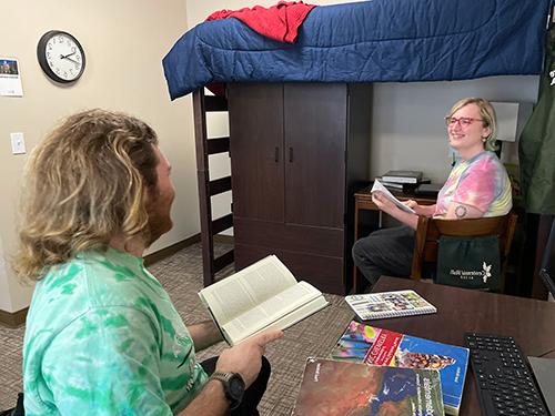 Two students in Centennial Triples room. A student has a book open in their hand and anotehr student is siting on a chair by the bunk bed